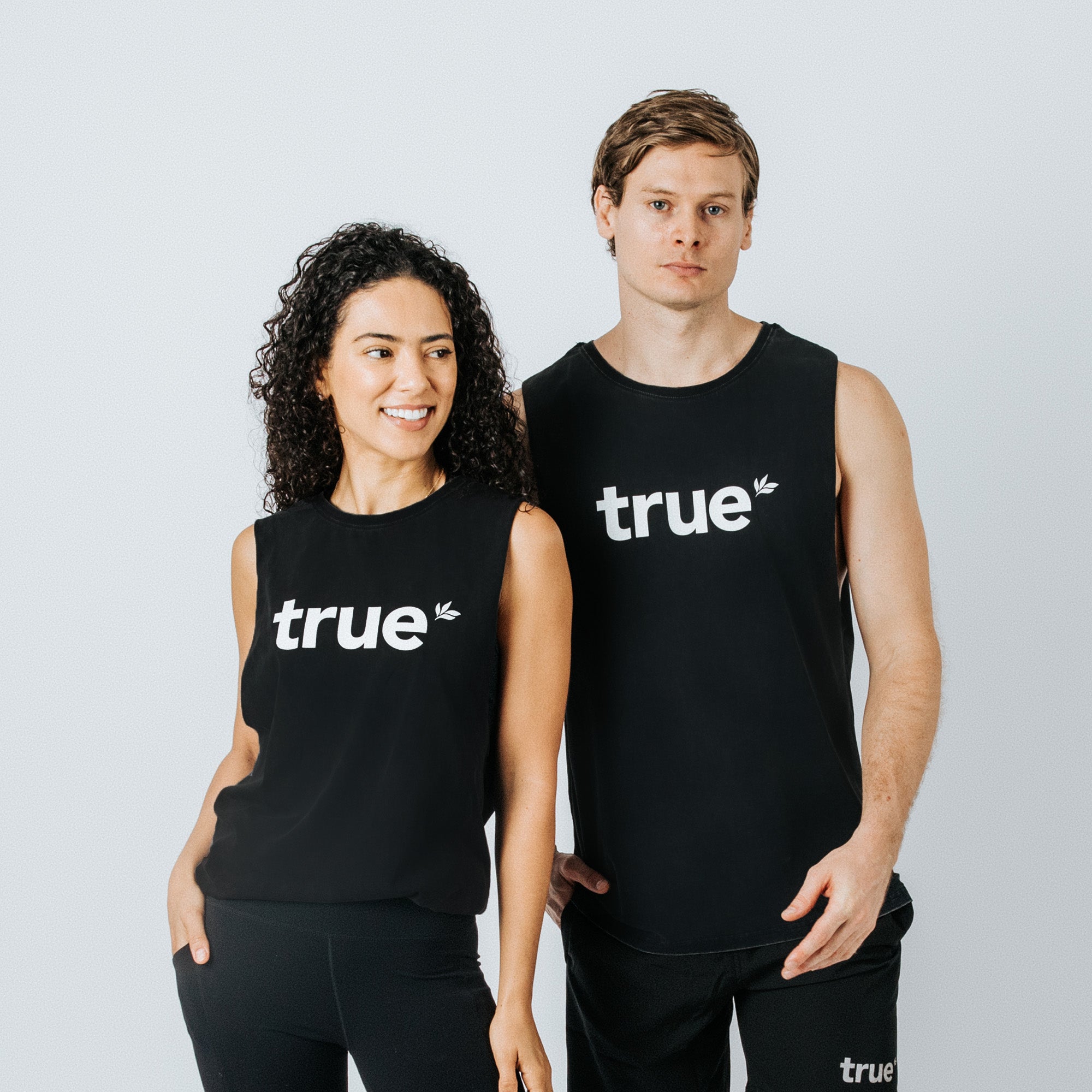 Black unisex sleeveless muscle tank top with white "true" logo, worn by male and female models in studio — black activewear tank, casual workout tee, branded fitness apparel.