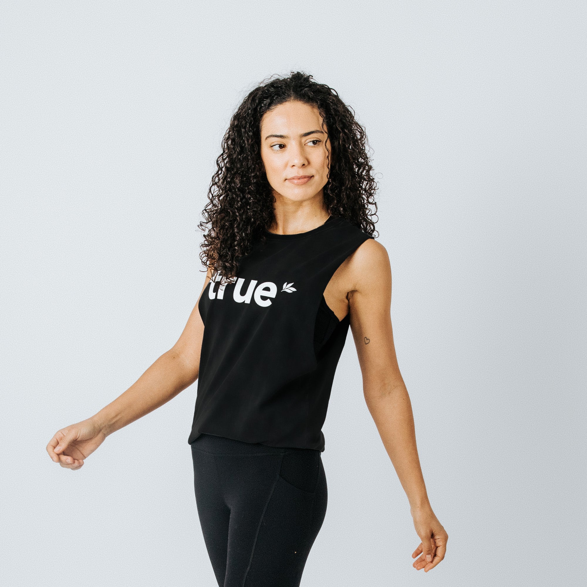 Women’s black muscle tank top with white "true" logo, athleisure yoga top styled with black leggings on a model against a light studio background — breathable workout tank for running, gym, and casual wear.