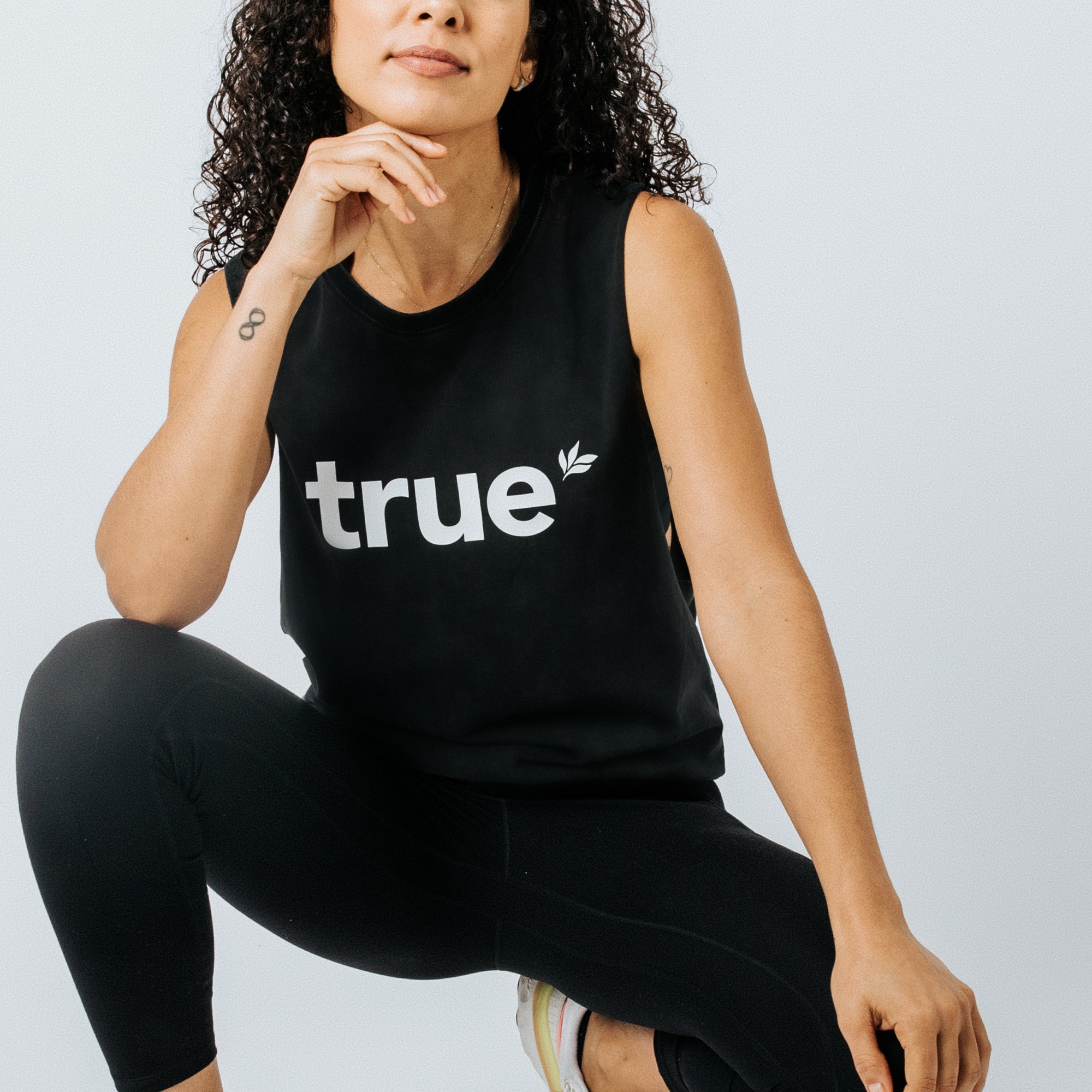 Woman wearing black "true" logo sleeveless tank top and black leggings, casual women’s activewear studio product shot