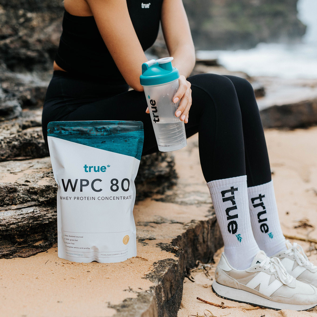 Woman sitting on rocky beach holding teal-lid True shaker bottle next to True WPC 80 whey protein concentrate pouch — fitness supplement product shot for beach workout lifestyle, Australia.