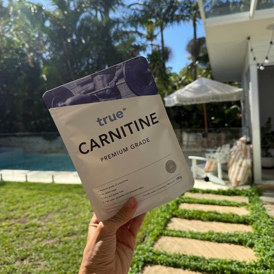 A hand holds a bag of true Carnitine Premium Grade supplement in an outdoor backyard setting with a swimming pool, palm trees, and a modern house in the background.