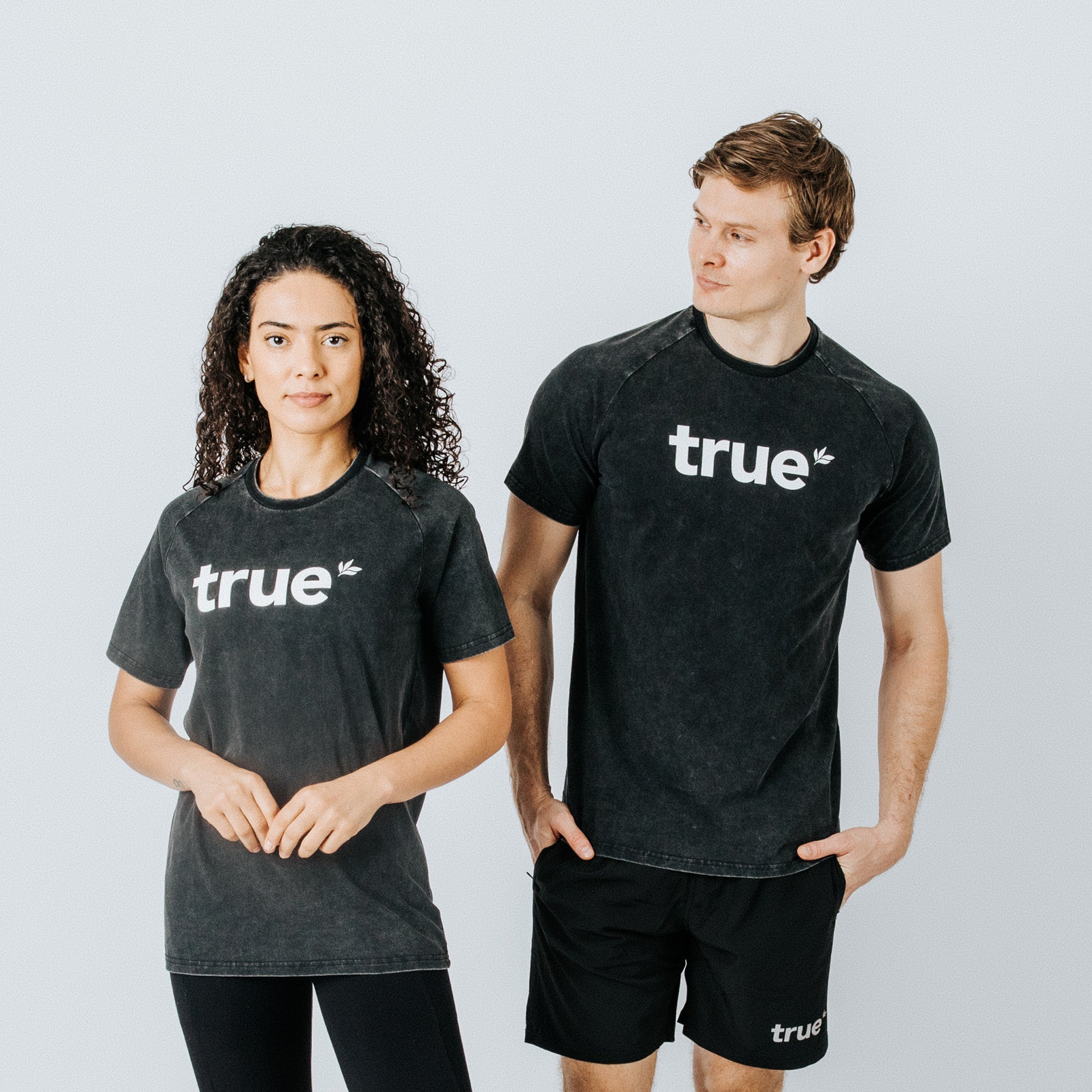 Two models (woman and man) wearing charcoal-washed "true" logo unisex cotton crewneck T-shirts, styled with black activewear shorts and leggings on light studio background — product image for online clothing store.
