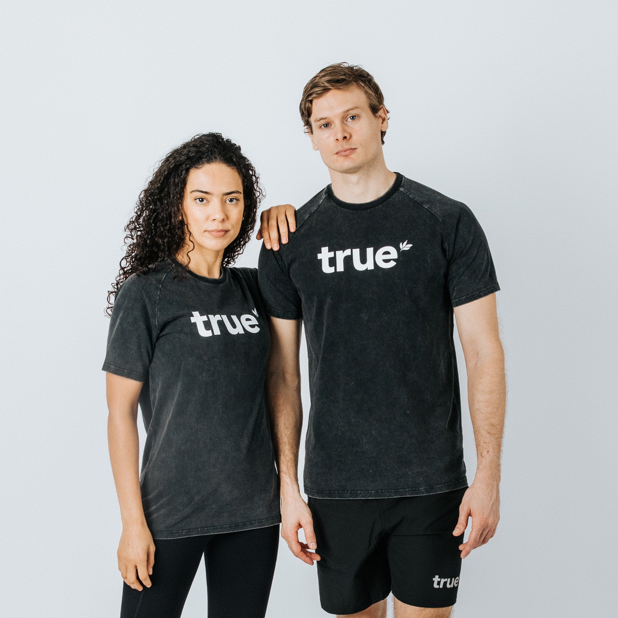 Two models (woman and man) wearing charcoal-wash unisex t-shirts with white "true" logo, studio shot on light gray background — charcoal tee, true brand, men&