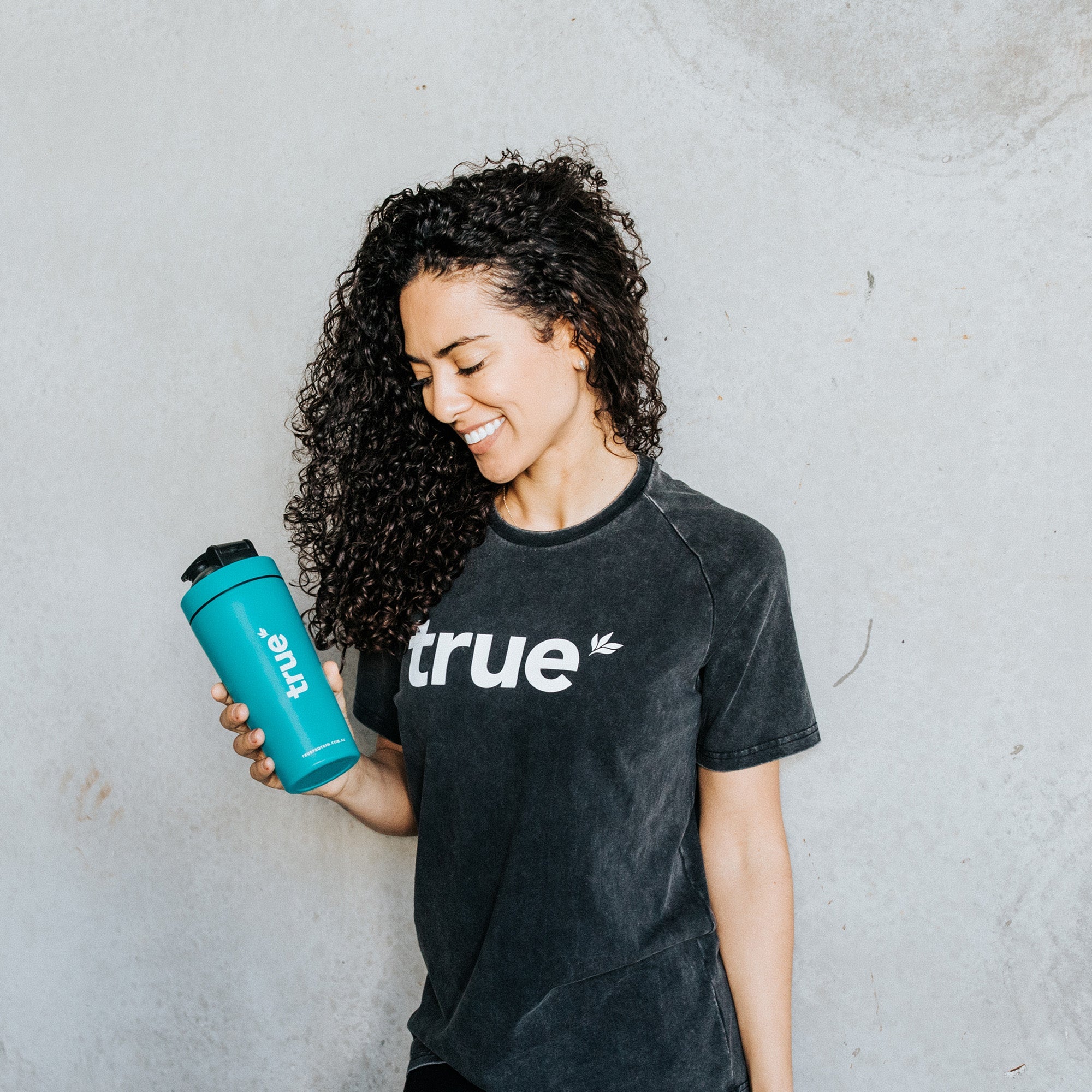 Woman holding teal "true" branded shaker bottle and wearing charcoal washed t‑shirt with white "true" logo, standing against light concrete background.