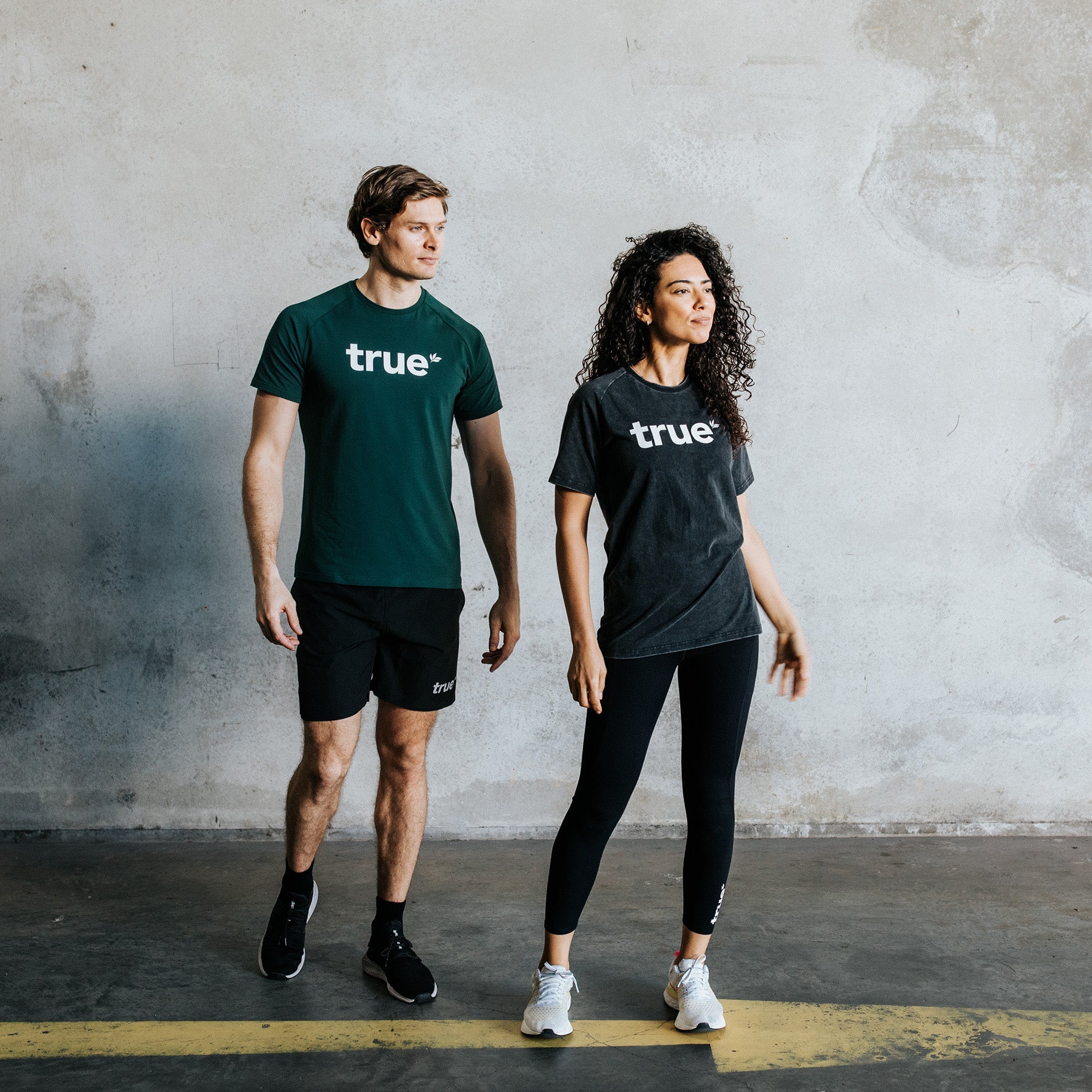 Male and female models wearing True branded charcoal black and forest-green t-shirts with black athletic leggings and shorts, posing in an industrial concrete studio — lifestyle activewear product photo.