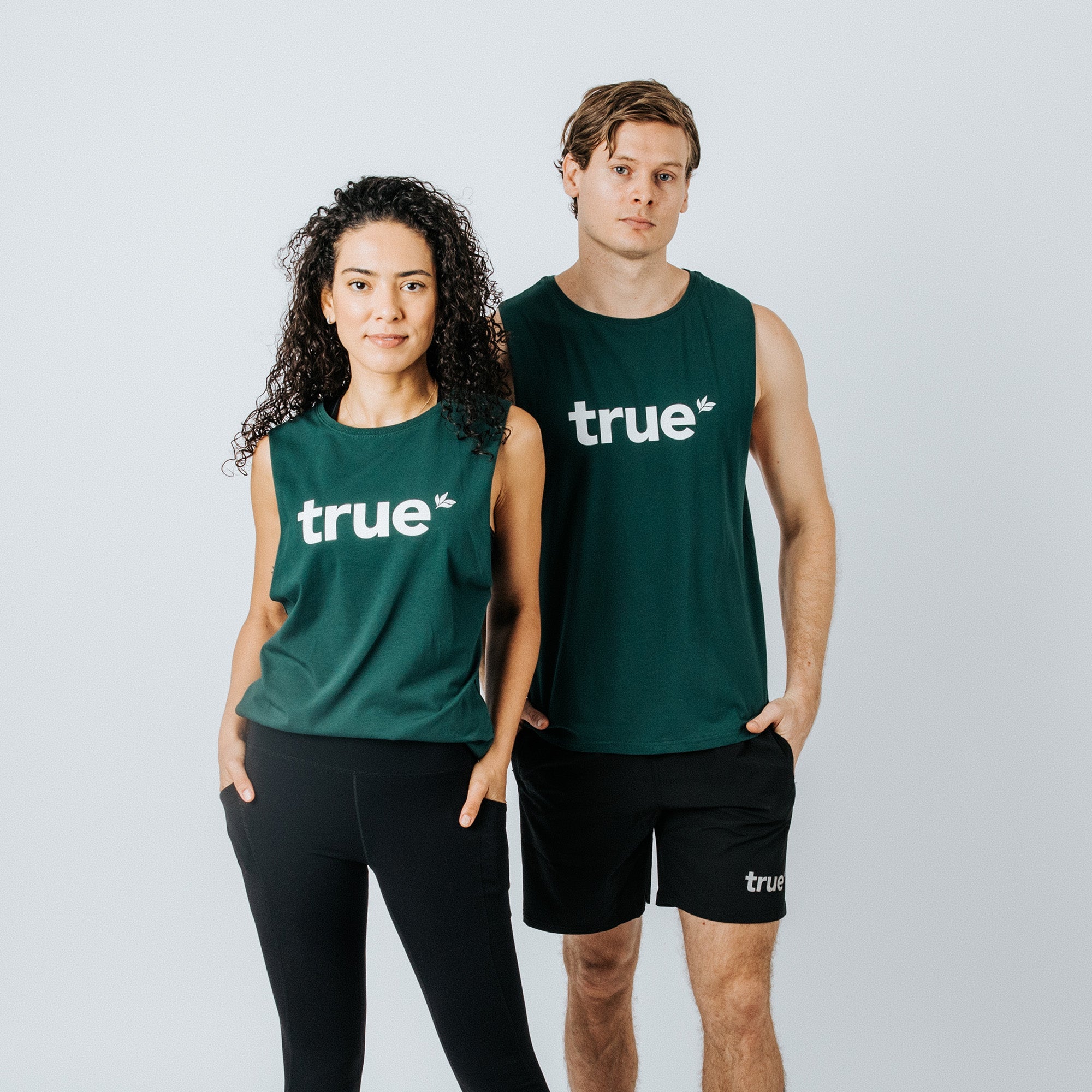 Two models wearing forest-green sleeveless "true" logo tank tops and black athletic bottoms, studio product shot showing front view of unisex fitness tanks for workout, sustainable activewear, apparel detail and fit.