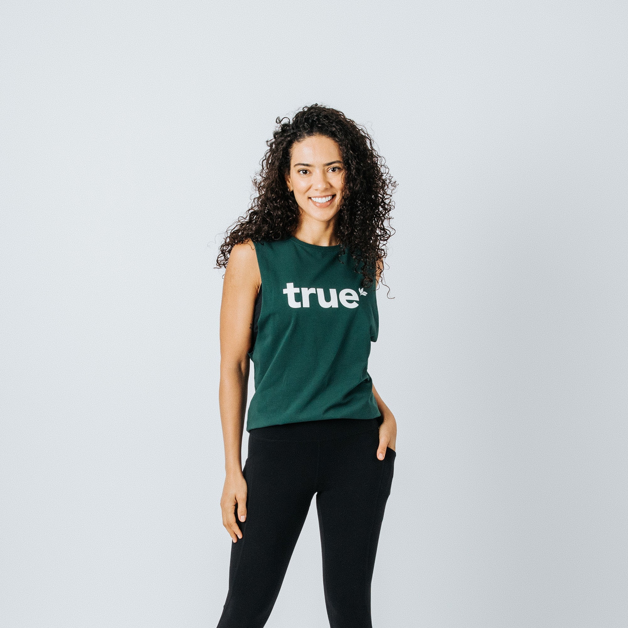 Woman wearing forest green "true" logo muscle tank top with black leggings — studio product photo for online activewear store.