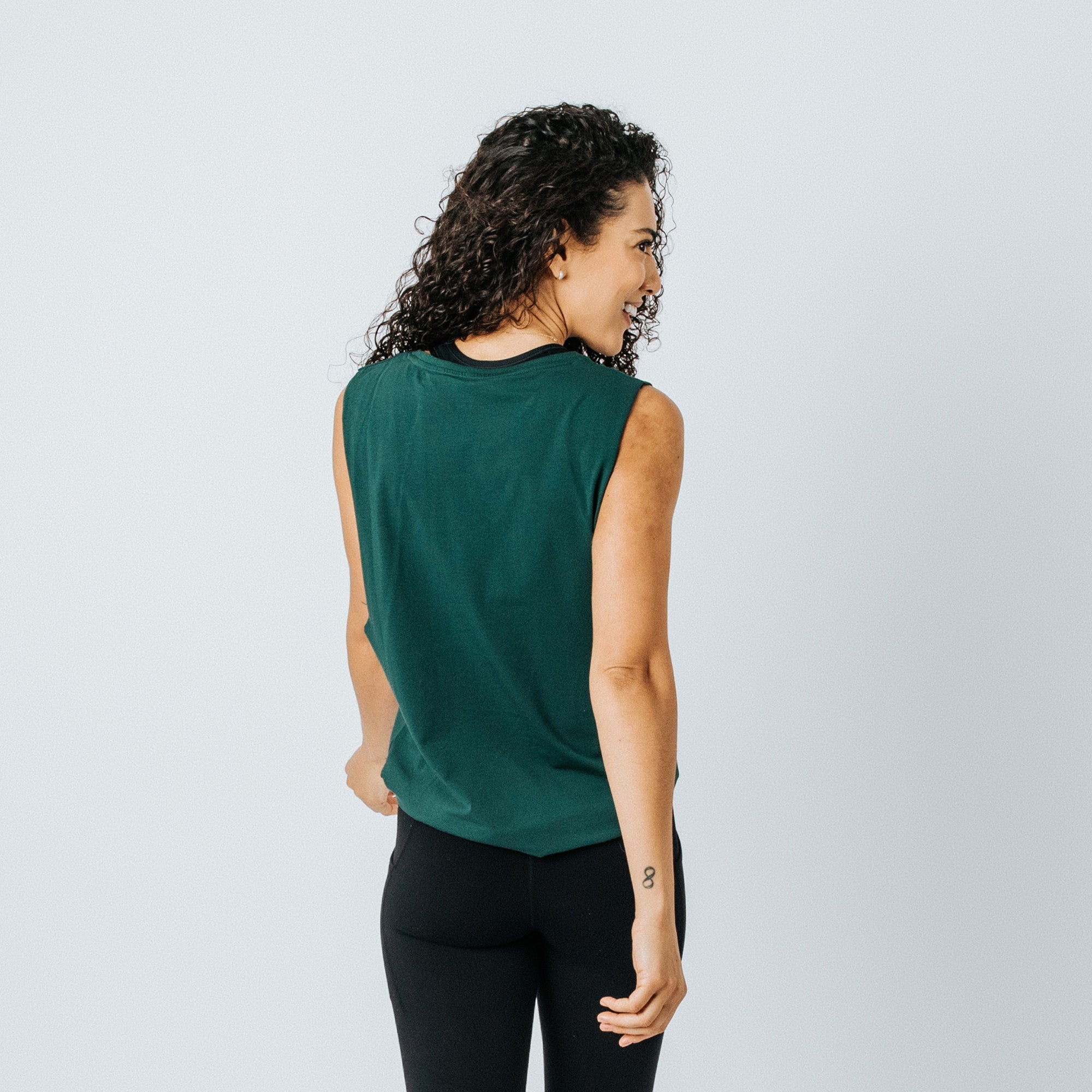 Woman wearing forest green sleeveless muscle tank top, back view on model, athletic fit paired with black leggings — studio product photo for women&