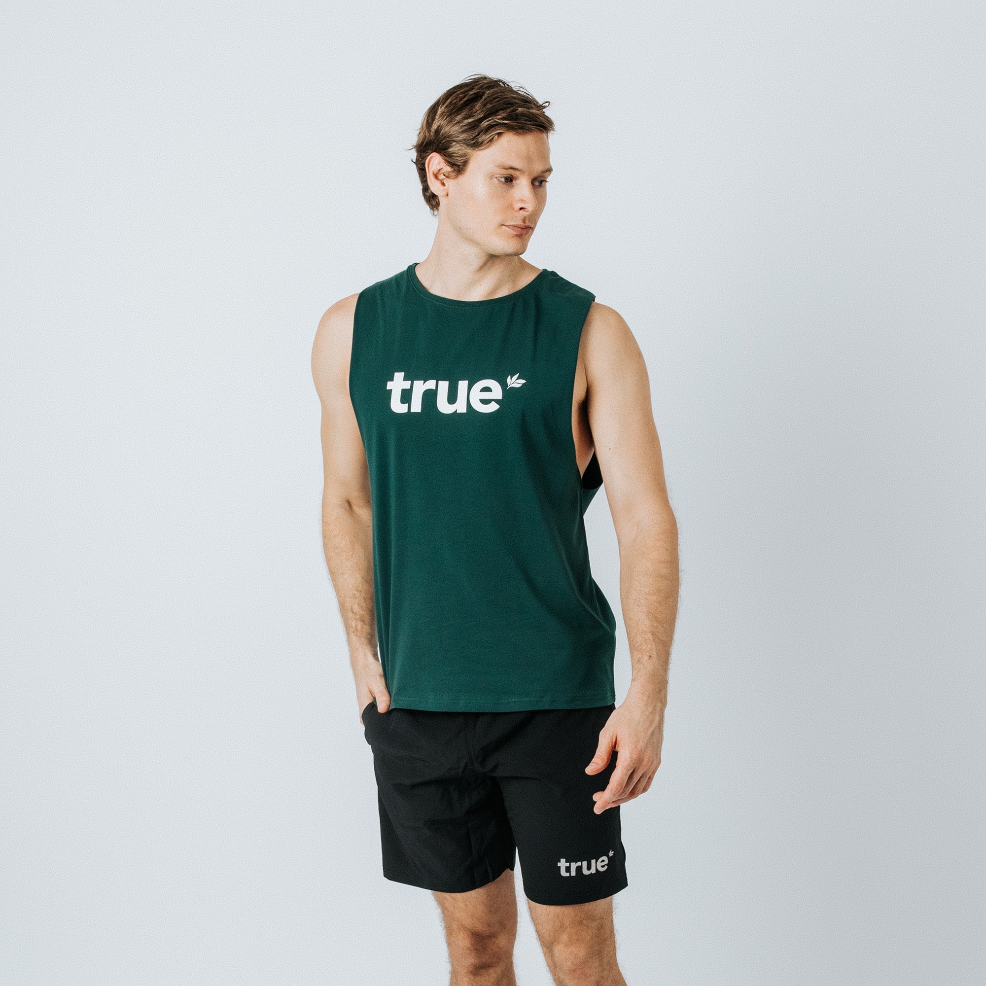 Male model wearing a forest-green "true" logo sleeveless tank top and black "true" logo athletic shorts, standing against a white studio background — men&