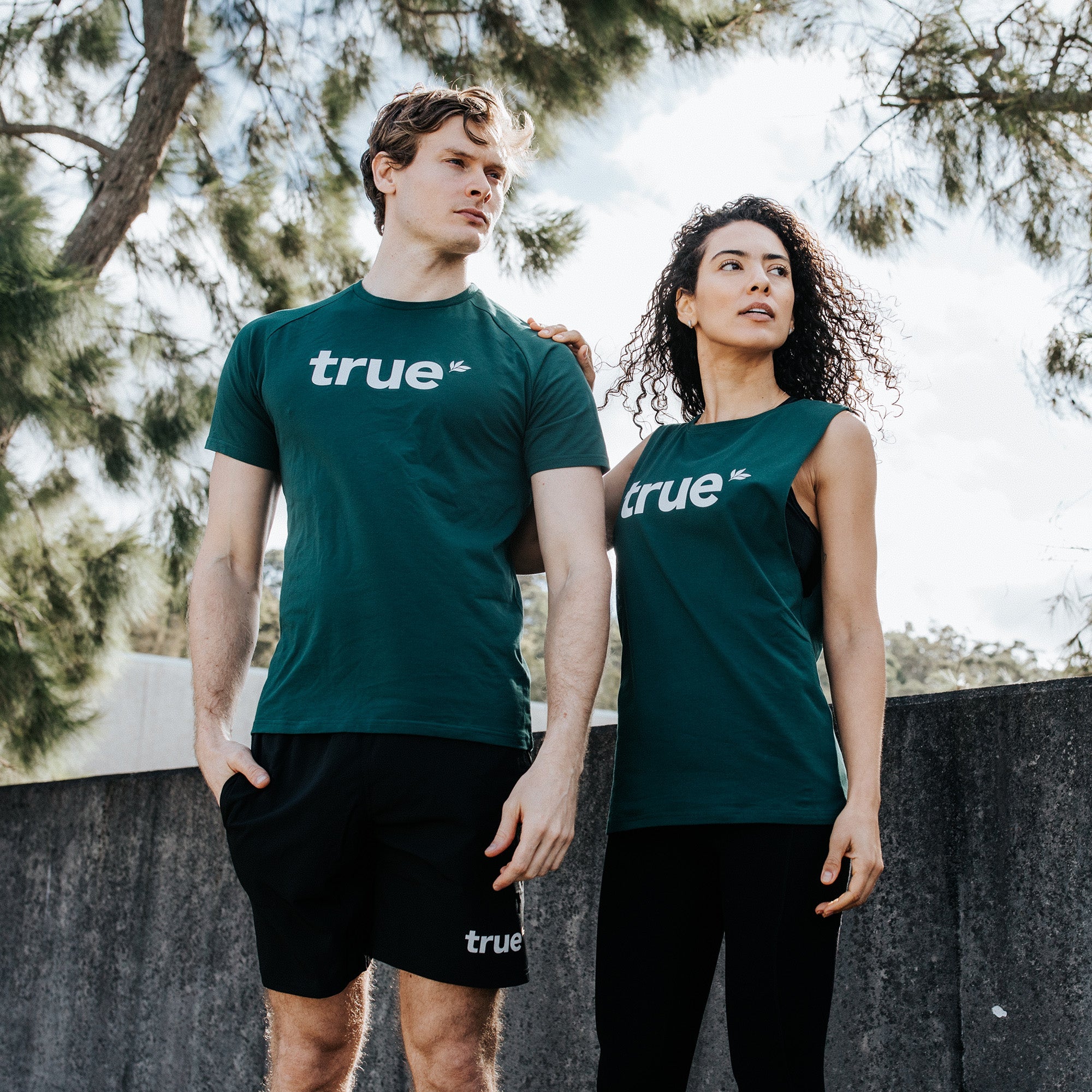 Two models wearing forest green "true" logo activewear — man in crewneck tee and black shorts, woman in sleeveless tank and leggings — posing outdoors by a concrete wall with pine trees and bright sky in the background.