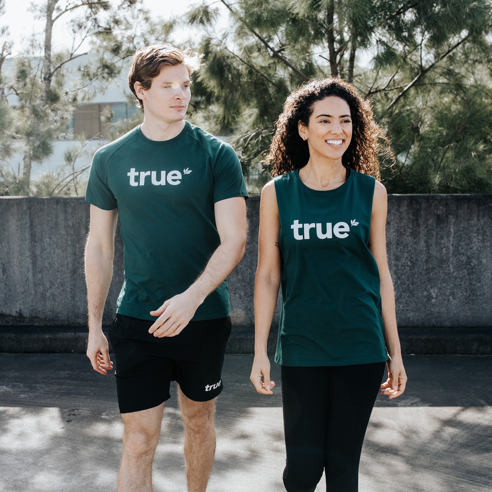 Two adults walking side-by-side in an outdoor urban setting wearing forest-green activewear: man in forest-green crew t-shirt and black shorts and woman in forest-green sleeveless tank, both with white "true" logo, casual fitness apparel, sunlight and trees in background.