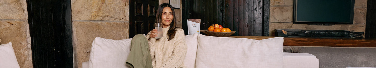 Woman in cream sweater sitting on light beige sofa holding a glass, lifestyle banner showing True ZMA supplement pouch and bowl of oranges on wooden shelf in a cozy neutral-toned living room.