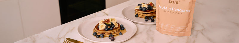 Protein Pancakes French vanilla pouch beside stacked protein pancakes topped with banana slices, blueberries and syrup on white plates on a marble kitchen countertop, gold fork visible.