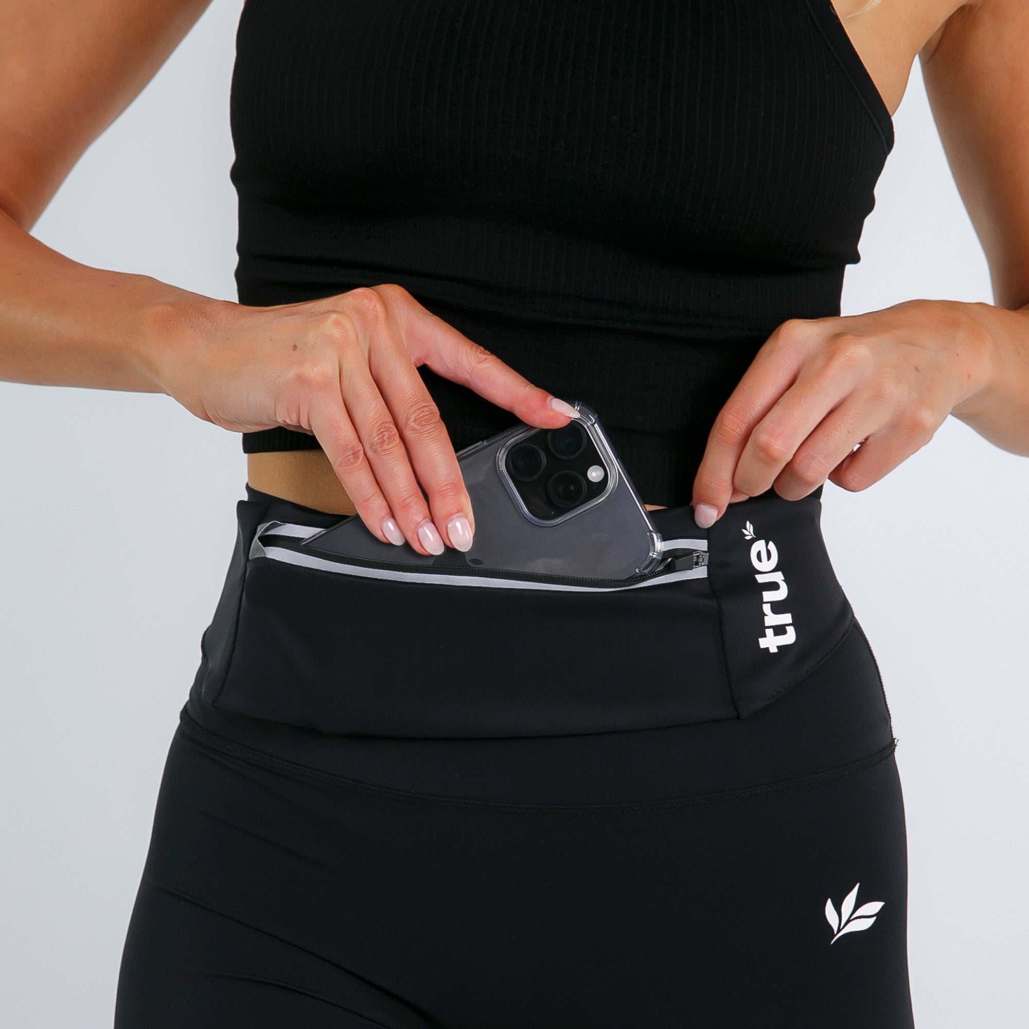 Woman wearing black running belt waist pack with zip pocket, inserting smartphone — women&