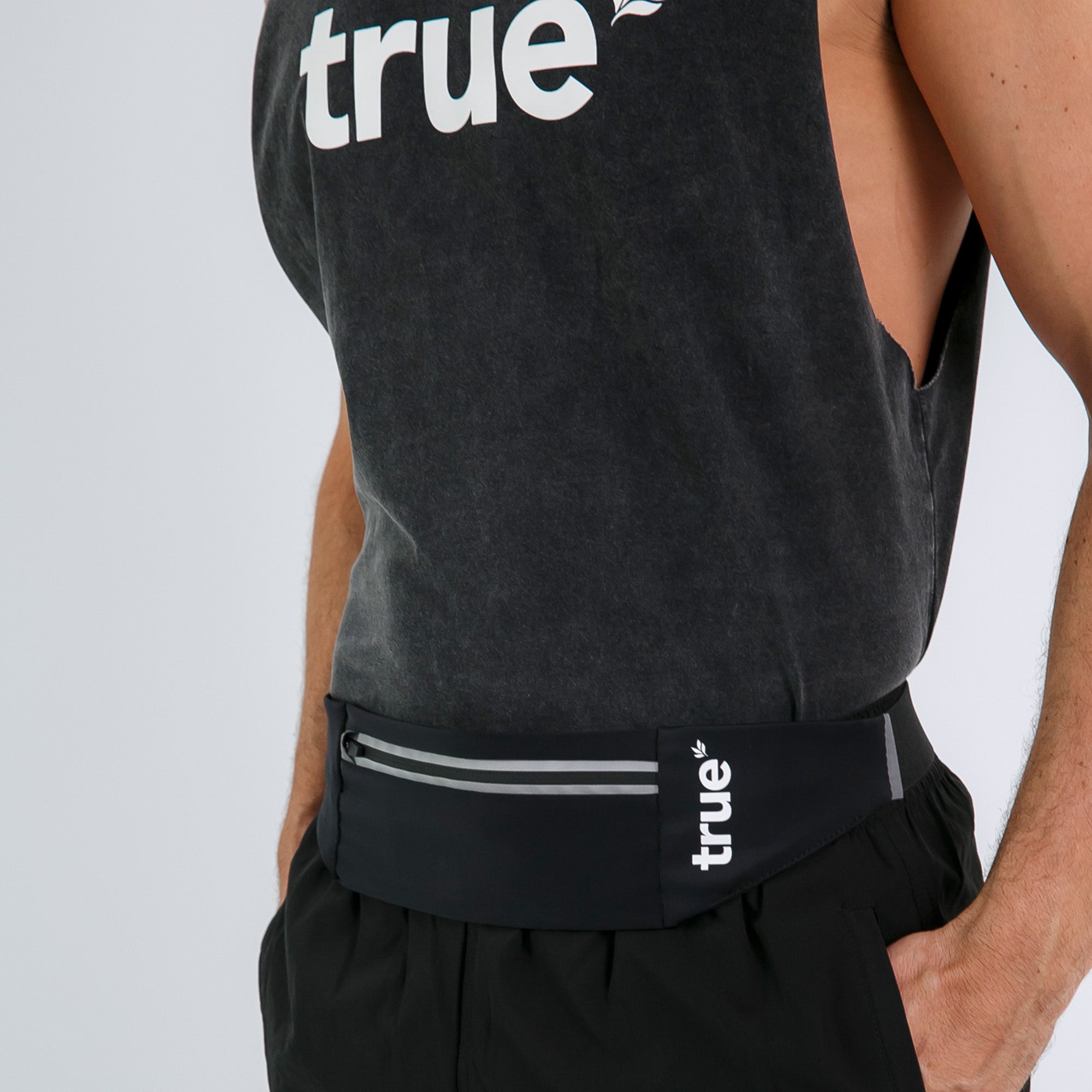 Man wearing black True branded running belt with reflective zipper pocket around waist, shown with black tank top and running shorts — athletic waist pack for hands-free running and travel.