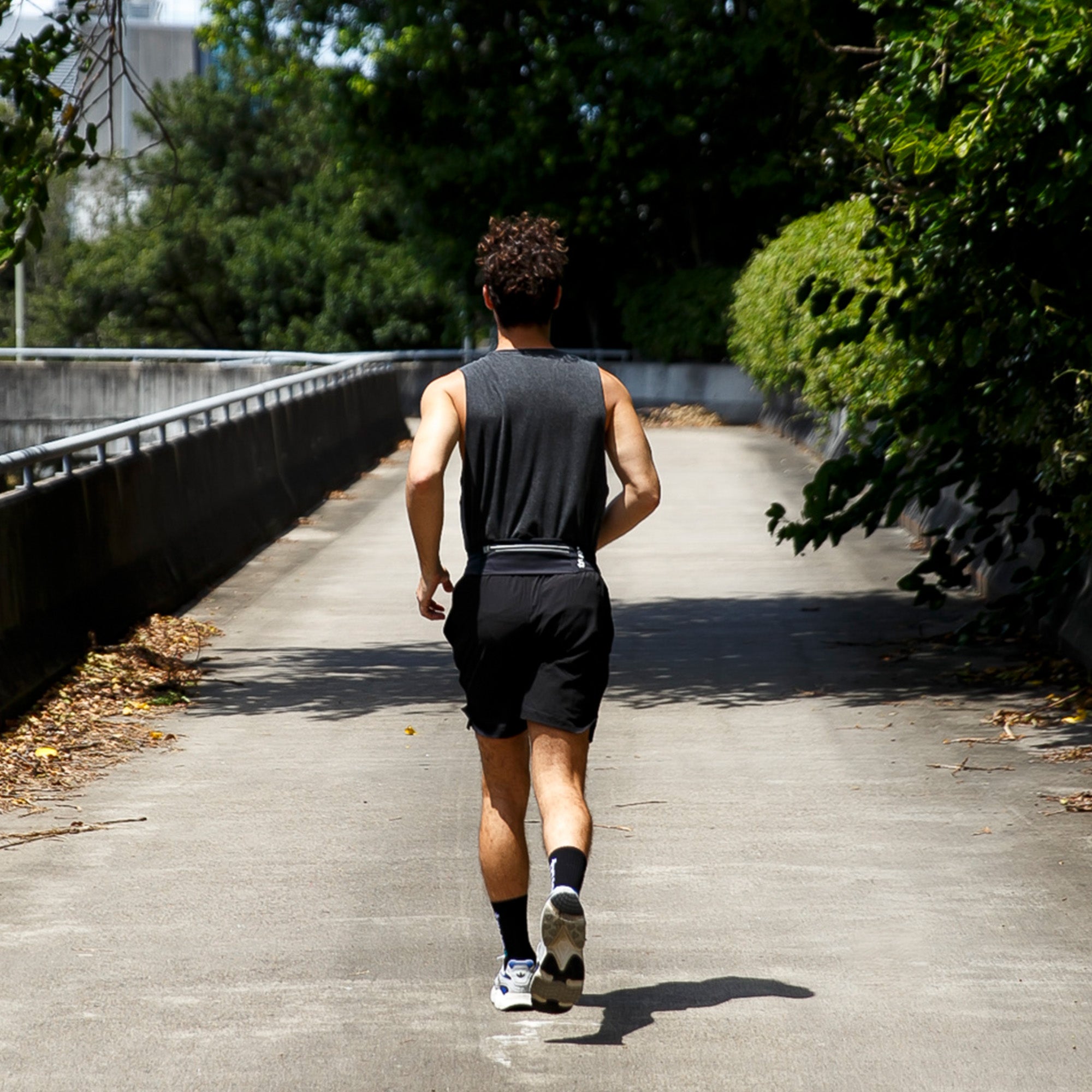 Man running away on urban tree-lined path wearing black shorts and a slim reflective running belt waist pack — men&