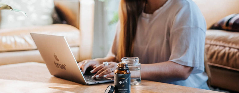 Open True Spark supplement bottle and cap on a coffee table next to a glass of water and a laptop with a person working from home in a cozy living room, lifestyle product shot.