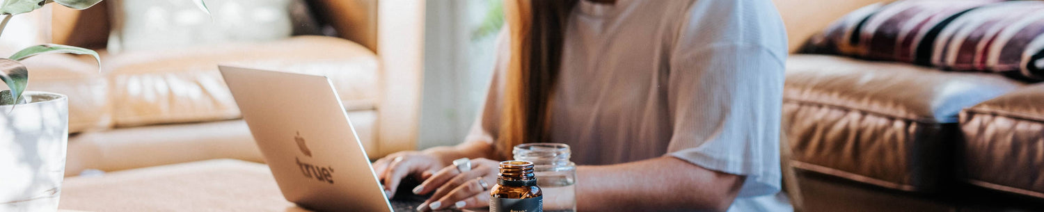 Woman working on laptop on living room floor with True-branded sticker, amber supplement bottle and glass of water nearby — work-from-home supplement lifestyle image