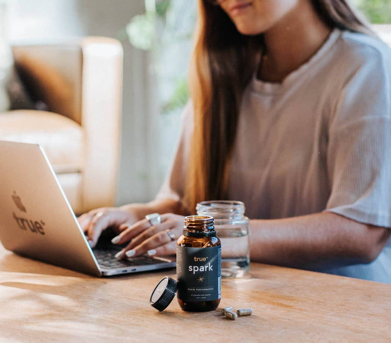 Woman working on laptop at wooden table in home office with open True Spark supplement bottle and capsules beside a glass of water, lifestyle product shot.