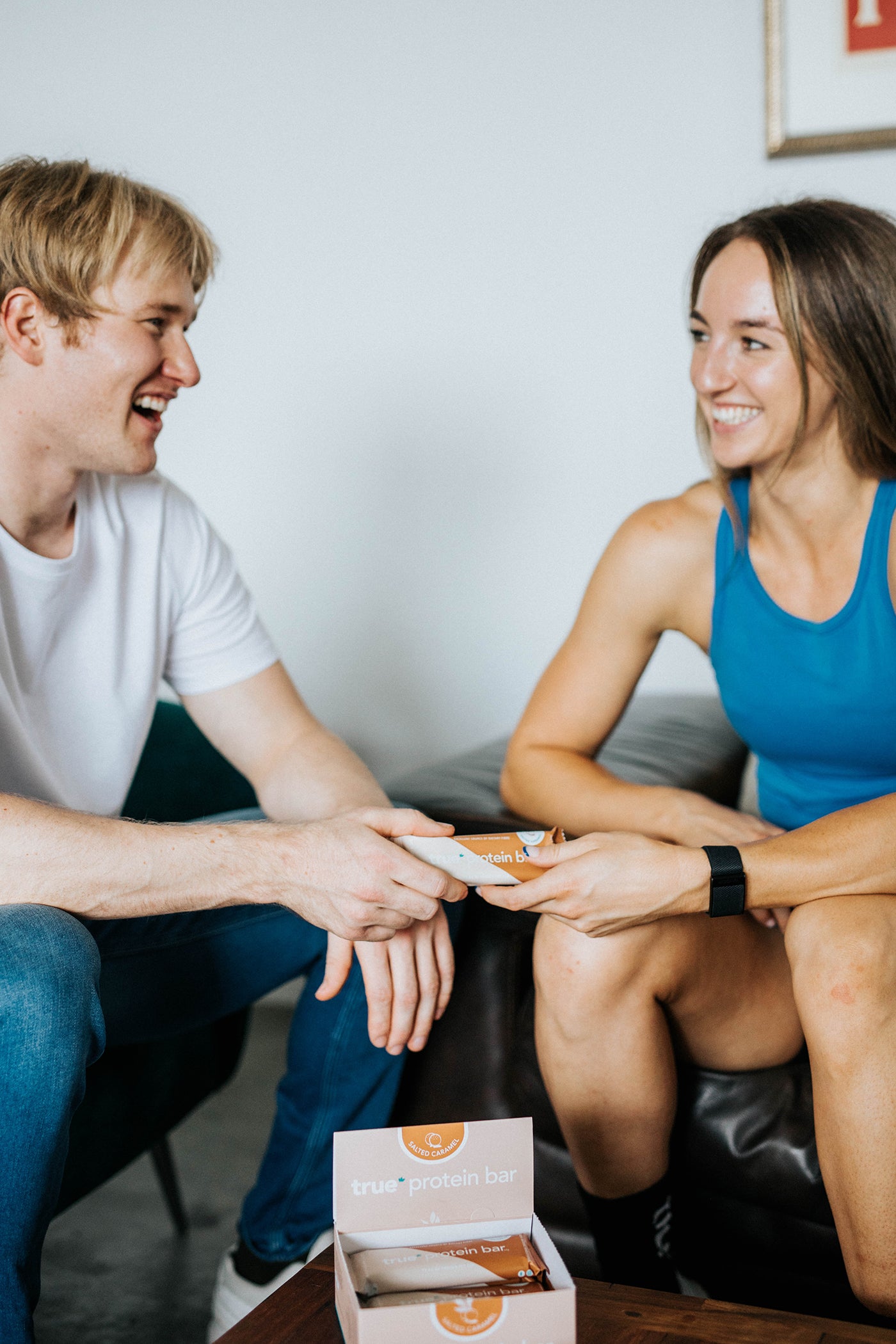 Two smiling adults in a living room passing a Salted Caramel True Protein bar, with an open box of True Protein bars on a coffee table—healthy snack, fitness nutrition product.