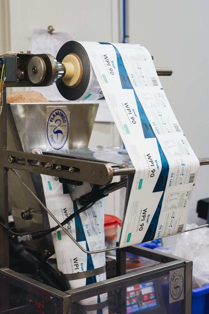 Packaging machine printing and rolling True Protein WPI 90 protein powder labels on a production line in a manufacturing facility