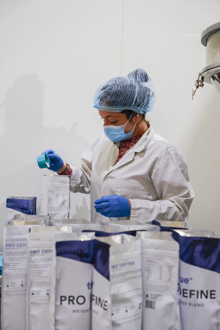 Worker in white lab coat, hairnet, mask and gloves measuring and filling True Protein "Pro Define" protein powder into packaging in a production facility, quality control and packaging process.