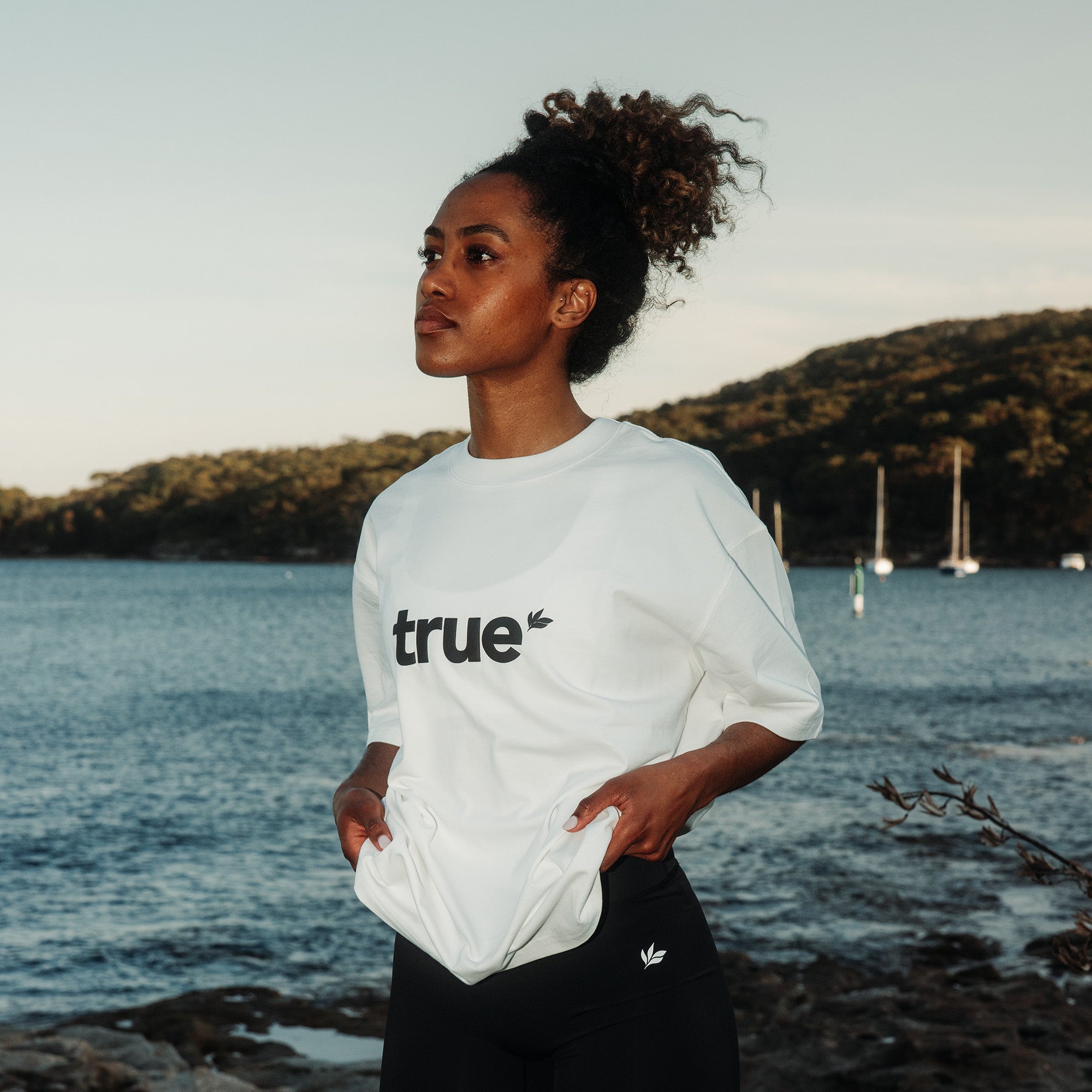 White unisex oversized tee with black "true" logo worn by a woman model standing at a coastal harbor with sailboats in the background — lifestyle product shot for casual eco-friendly clothing.