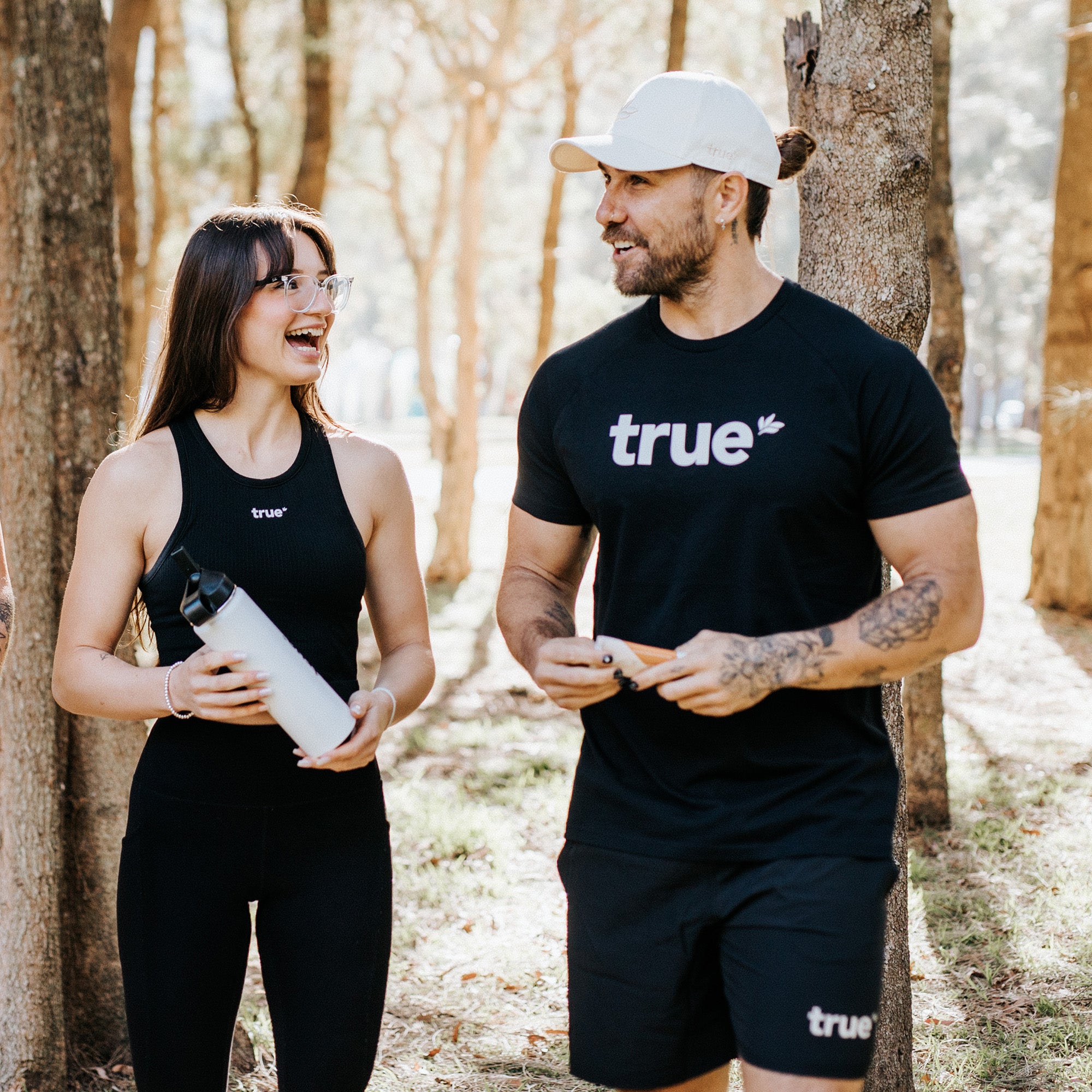 Two fit adults in a sunlit park wearing black "true" activewear; woman holding a white stainless water bottle and smiling, man in cap and shorts with tattooed arms — lifestyle fitness product shot for workout gear and water bottle.