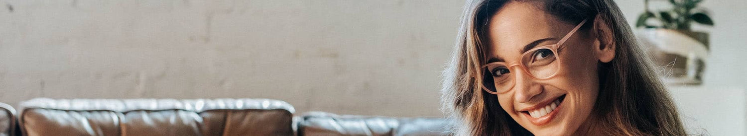 Smiling young woman wearing clear-framed glasses seated on a brown leather sofa in a bright modern loft-style living room with a potted plant in the background