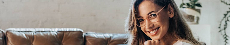 Young woman wearing glasses smiling and using a tablet on a leather sofa in a bright home office — remote work and online learning concept
