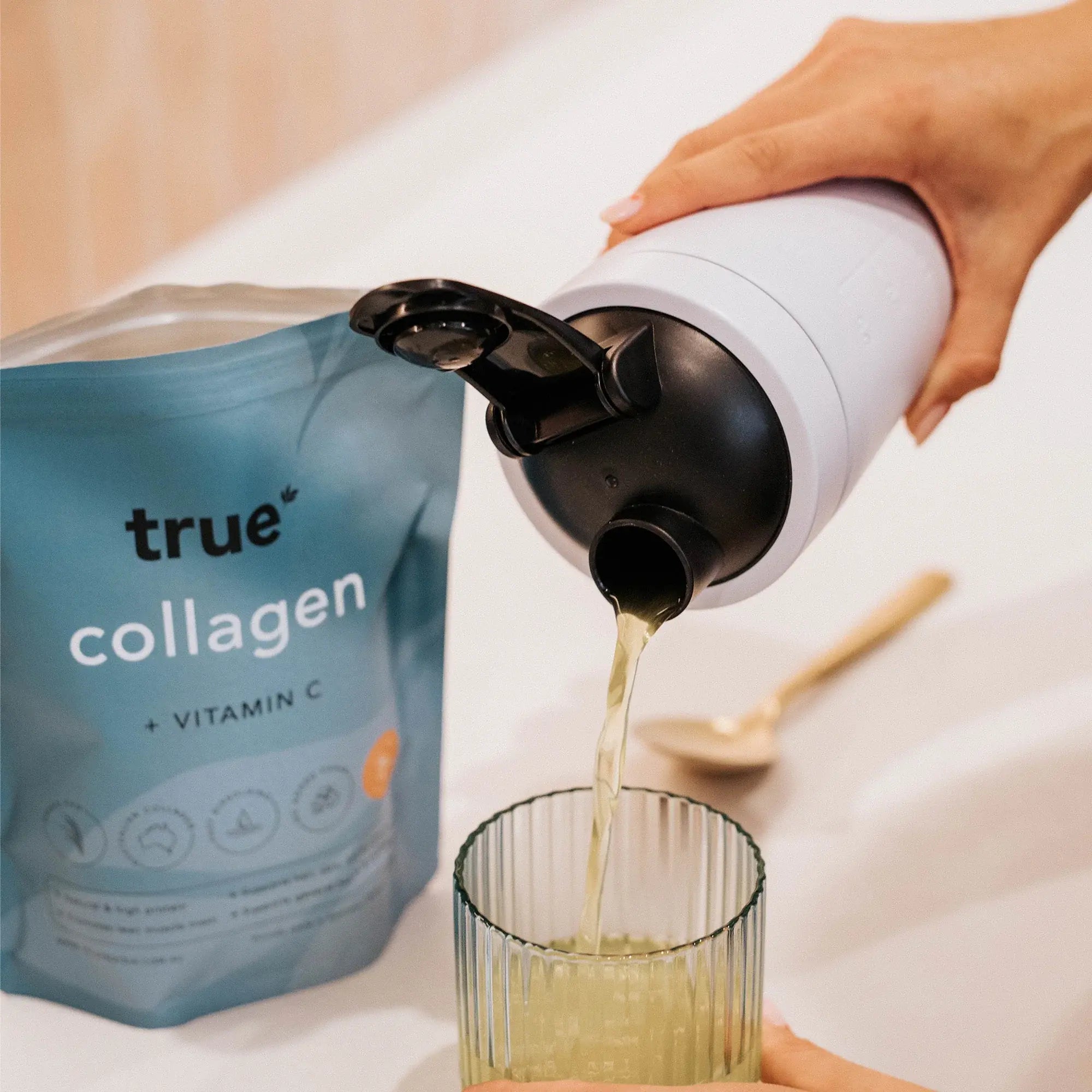 True Collagen + Vitamin C powder pouch next to a shaker bottle pouring pale yellow collagen drink into a ribbed glass — collagen powder supplement, collagen drink, vitamin C beverage.