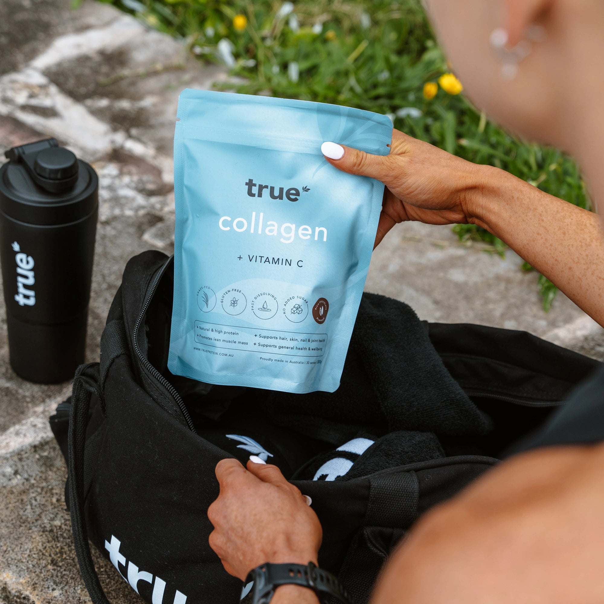 True Collagen + Vitamin C powder pouch (True brand) held above a black gym bag with matching True shaker bottle outdoors — fitness supplement product photo, Australia-focused SEO.