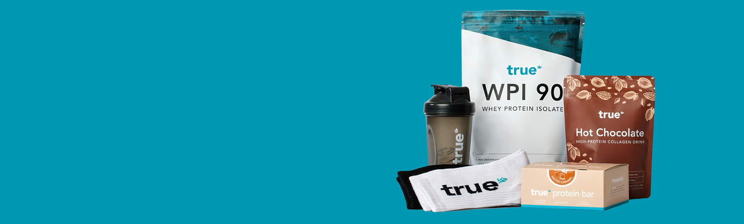 True Nutrition fitness bundle: True WPI 90 whey protein isolate pouch, True Hot Chocolate collagen drink pouch, True protein bar box, branded shaker bottle and socks on teal background — gym supplement gift pack for muscle recovery and protein users.