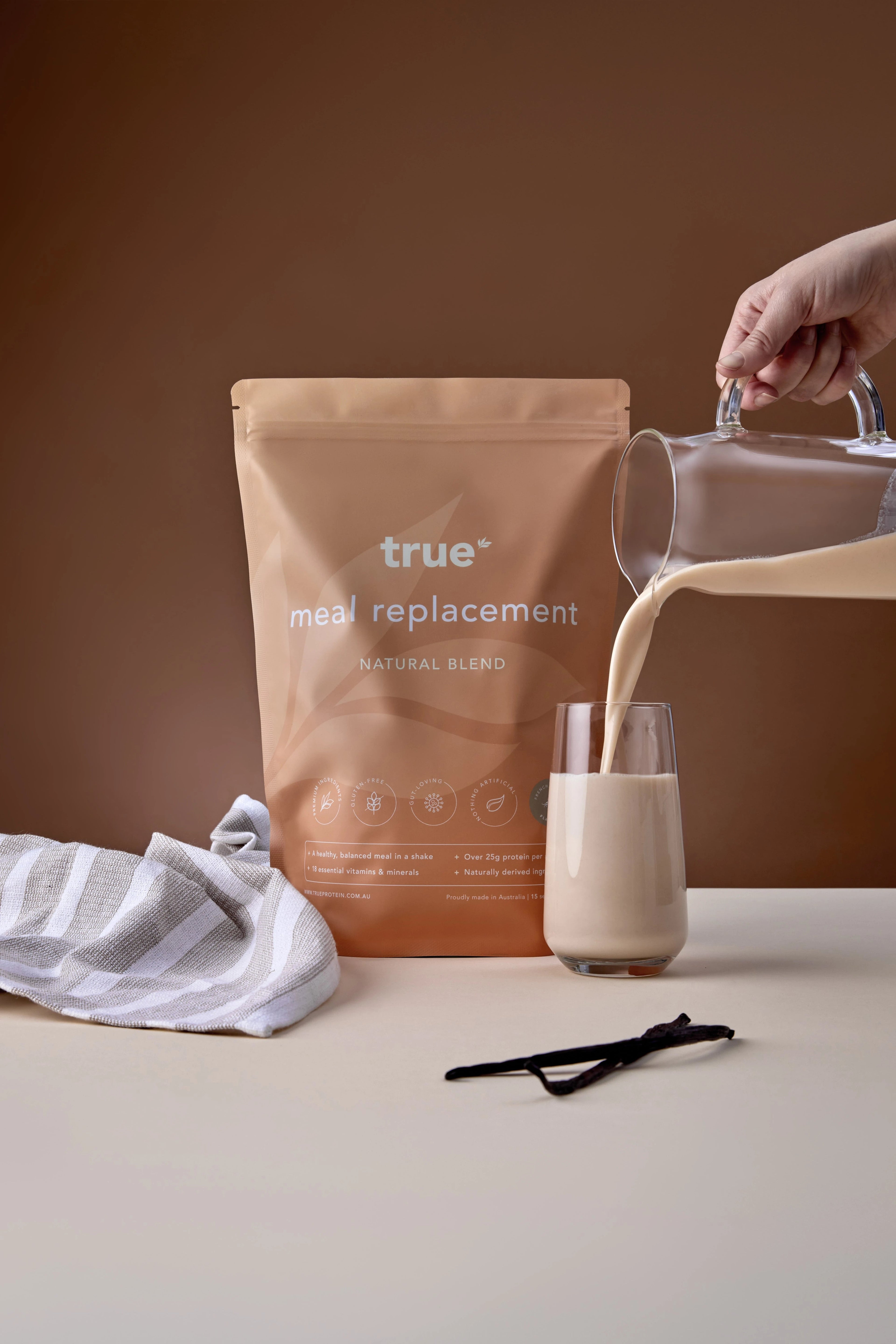 True Protein French Vanilla meal replacement pouch with a hand pouring creamy vanilla shake from a glass pitcher into a tall glass, vanilla pods and striped cloth on table, natural blend packaging.