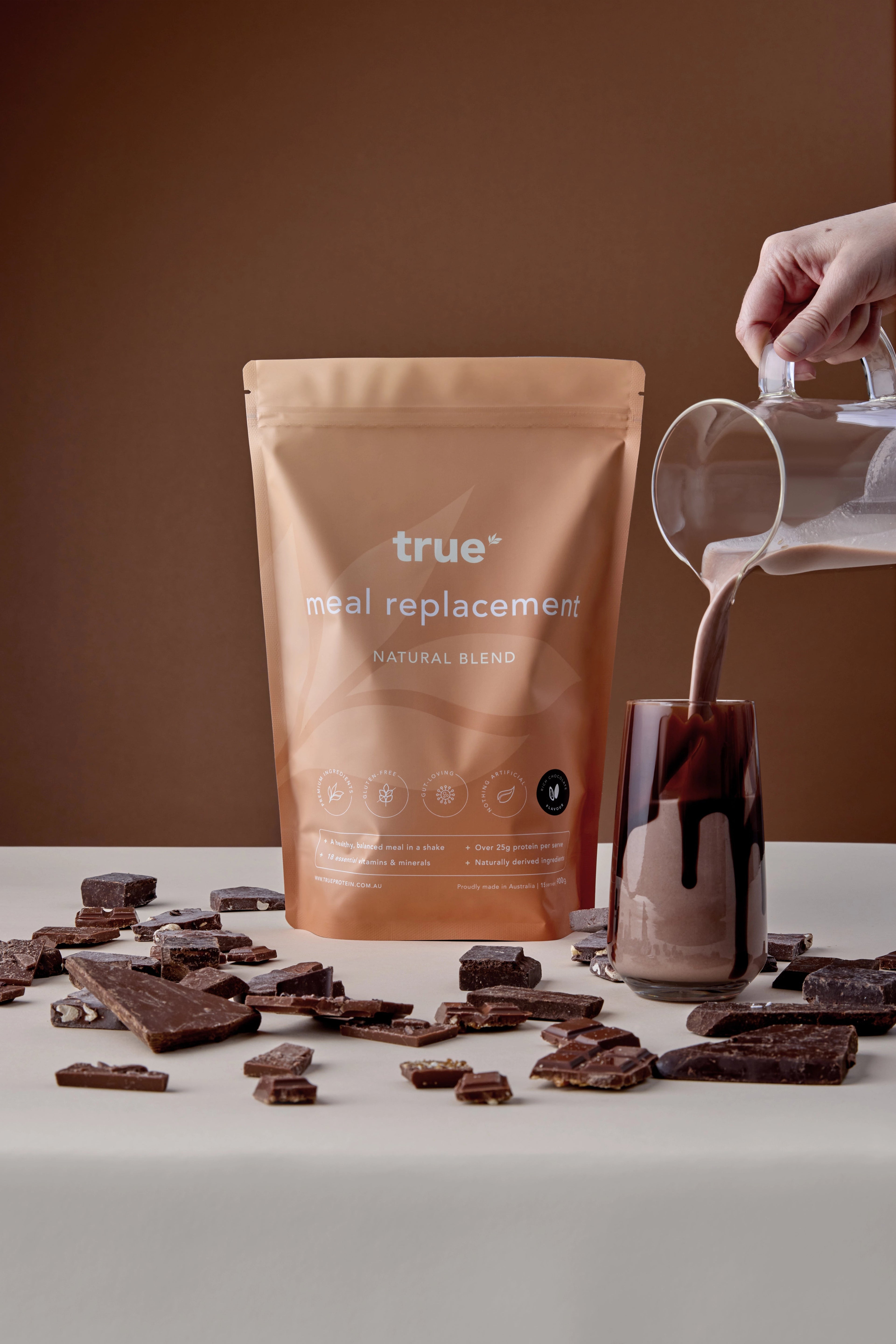 True meal replacement natural blend rich chocolate powder pouch with chocolate shake being poured into glass and scattered chocolate pieces — Australian-made product.