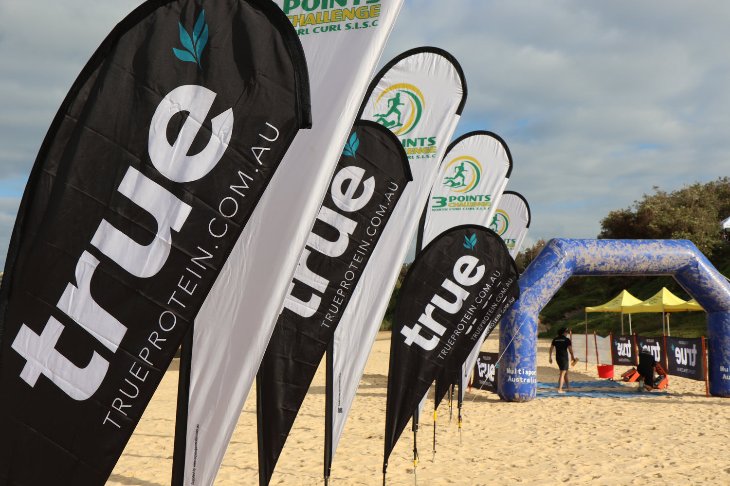 Row of black and white True Protein teardrop flags and event banners on the sand at a beach sports event, North Curl Curl Beach, Sydney, Australia