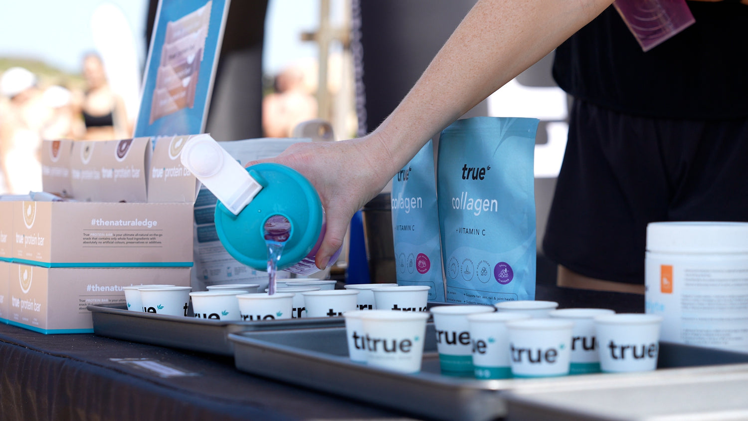 True Collagen sample tasting station: hand pouring purple collagen drink into branded "true" sample cups on trays beside True protein bar boxes at an outdoor fitness event.