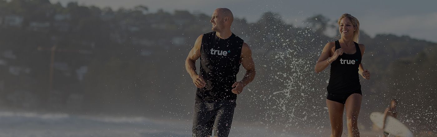 Fit man and woman running and splashing on an ocean beach at sunrise wearing "true" tank tops — active lifestyle, health supplement brand imagery.
