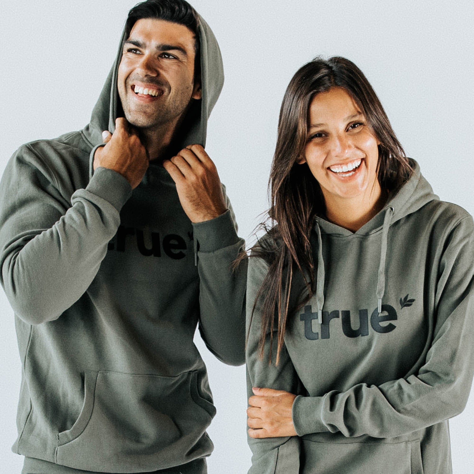 Two smiling models (man and woman) wearing khaki unisex hoodies with black "true" logo on front — cozy urban streetwear pullover with kangaroo pocket, drawstring hood, relaxed fit.