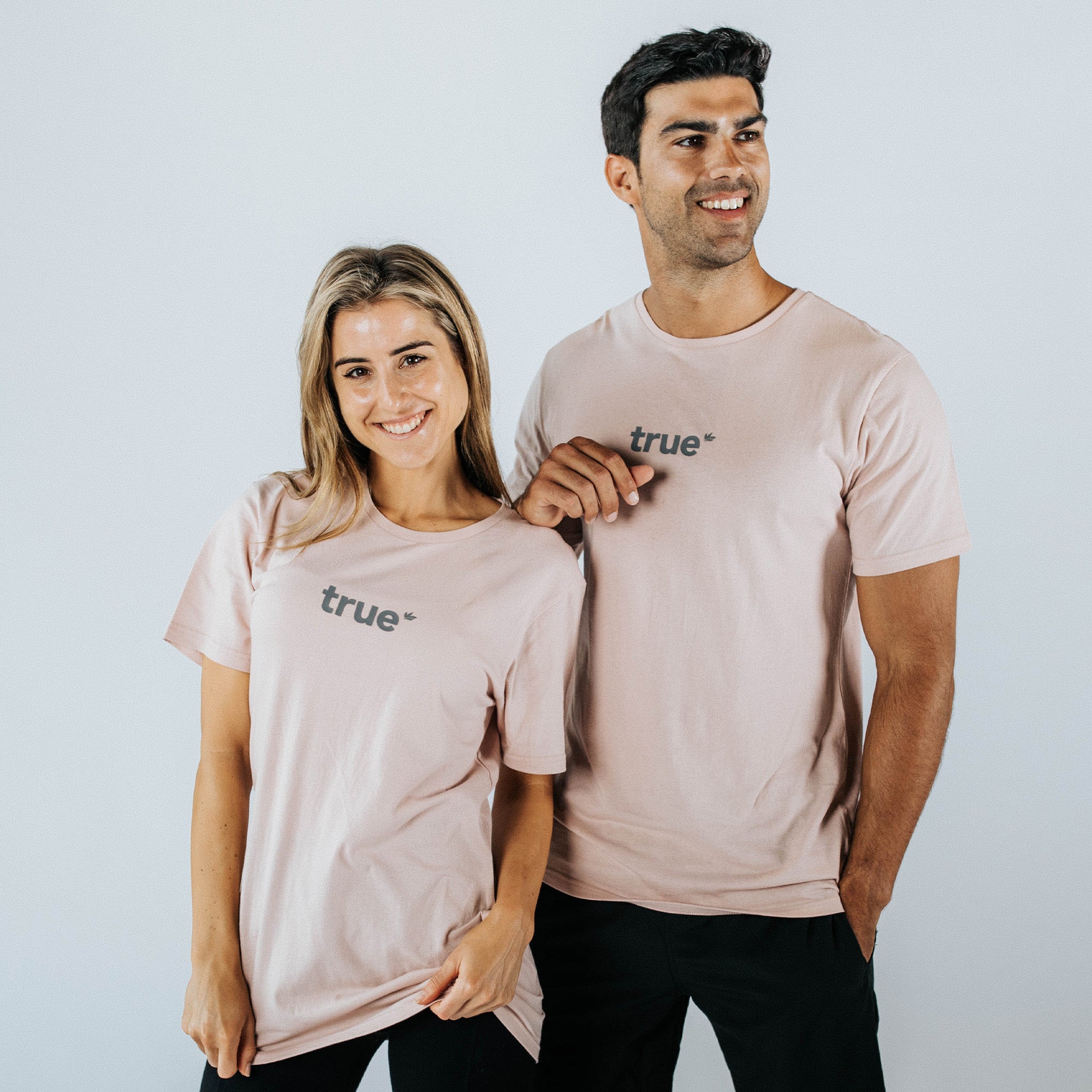 Studio product photo of male and female models wearing matching pale pink unisex "true" logo cotton short-sleeve t-shirts, casual lifestyle fit for men and women — ecommerce apparel.