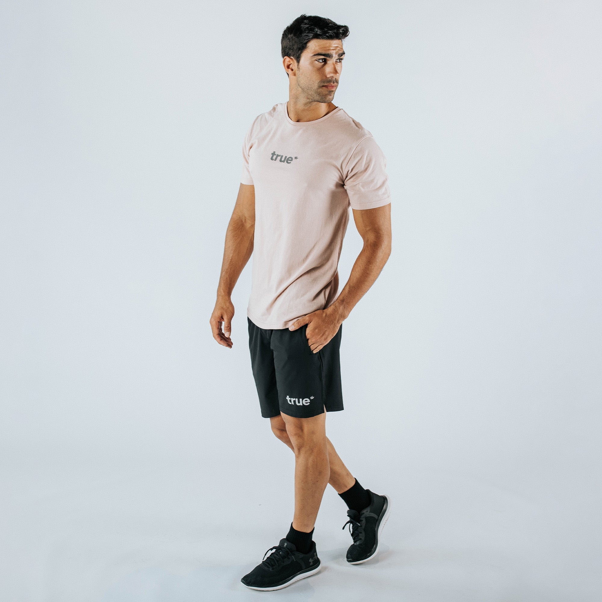 Male model wearing dusty pink "true" crew-neck t-shirt and black "true" athletic shorts with black running shoes, studio product photo on white background.