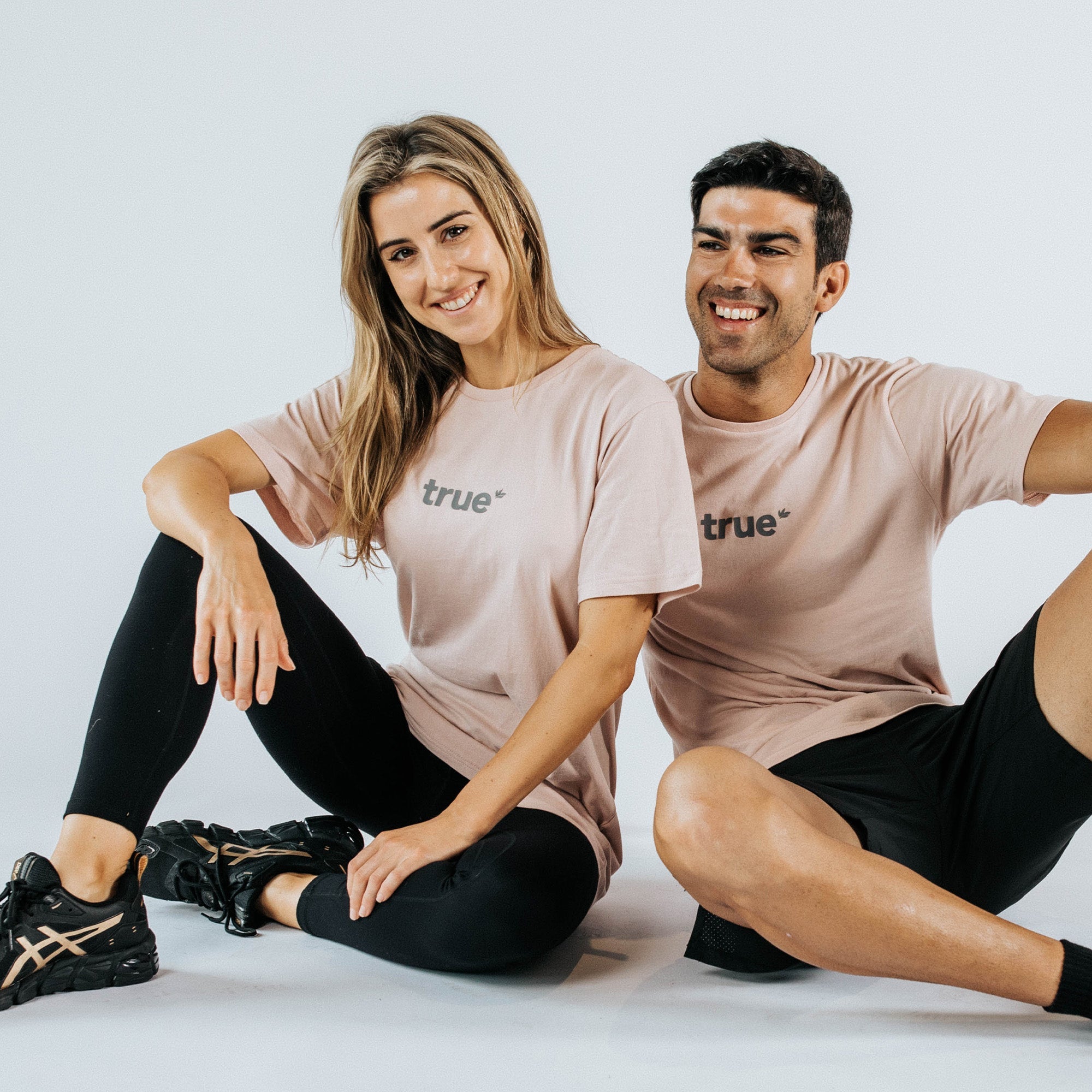 Two models sitting on a white studio floor wearing dusty pink "true" graphic t-shirts with black leggings and shorts — casual unisex tee product photo for online clothing store.
