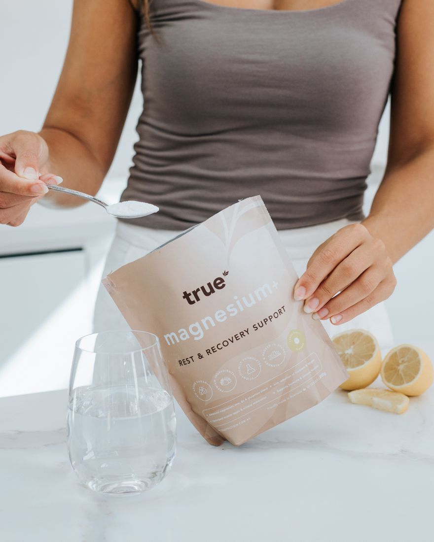 Woman scooping True Magnesium+ powder from a 250g pouch beside a glass of water
