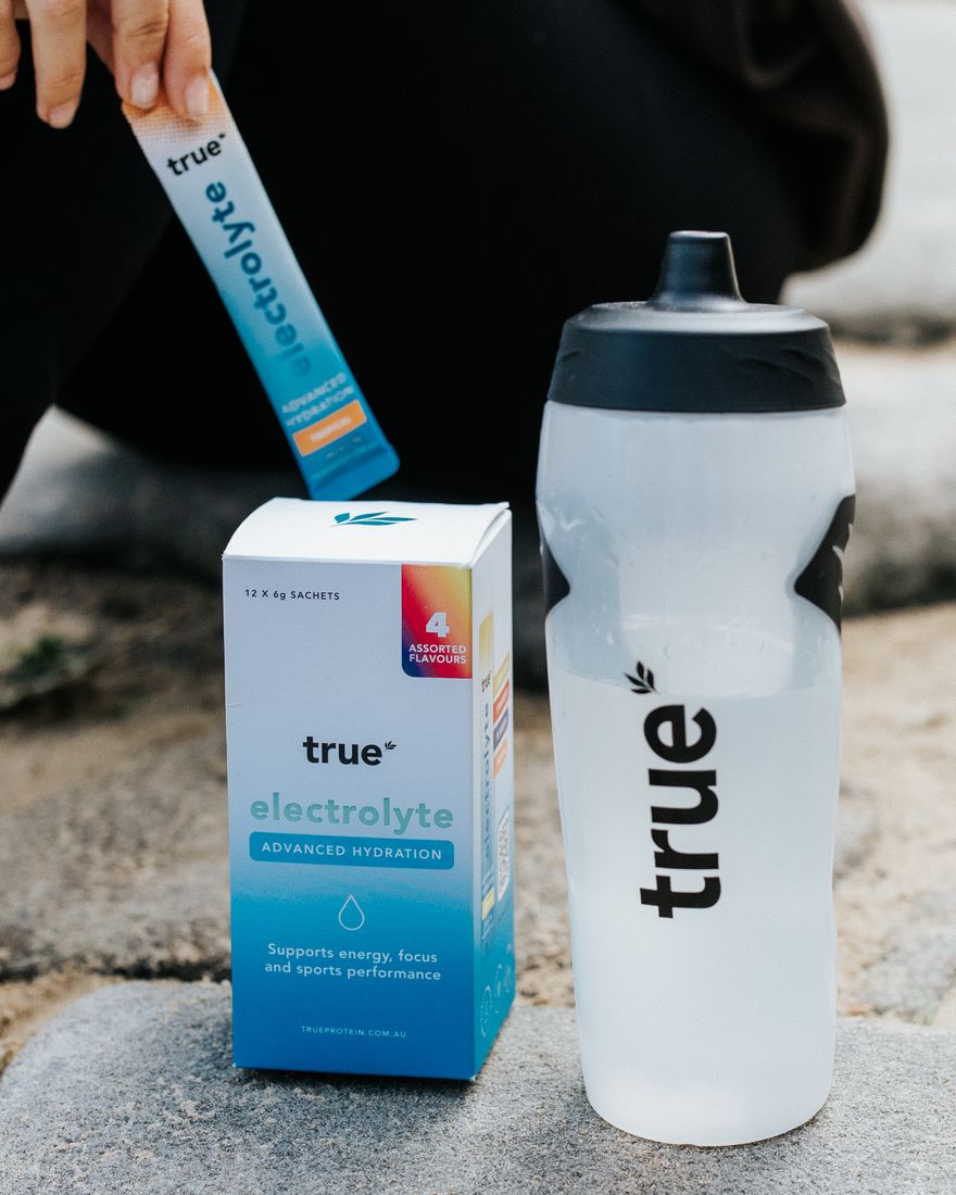 A True Electrolyte sachet being pulled from the Travel Pack box beside a True drink bottle on a sandy rock