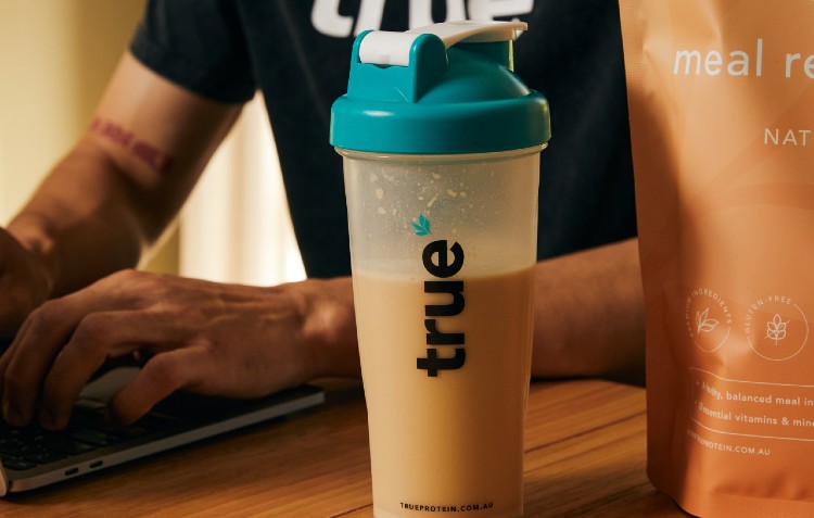True Protein shaker filled with a meal replacement shake on a desk next to a person working on a laptop, illustrating a healthy nutrition choice to support a balanced lifestyle during screen time.