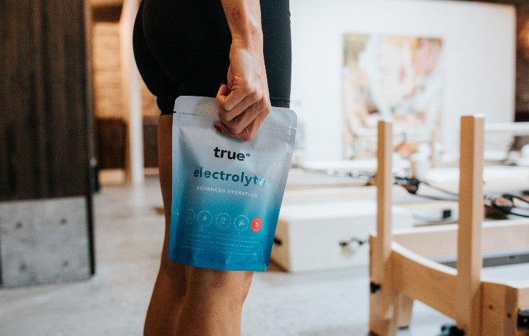 A person in athletic clothing holds a pouch of True Electrolyte Advanced Hydration in a fitness studio.