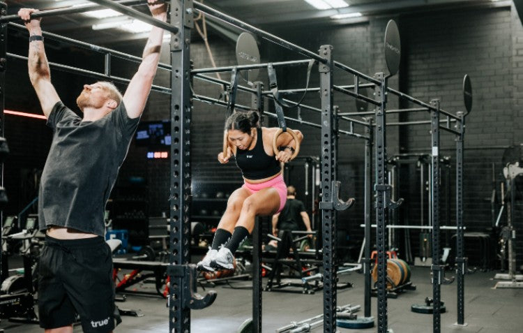Two athletes focus on a high-intensity functional fitness workout in a dedicated strength training gym, supported by True Protein. A man hangs from a pull-up rig while a woman performs a muscle-up on gymnastics rings.