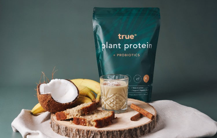 True Protein Plant Protein + Probiotics package displayed with a banana bread smoothie, fresh fruit, and baked goods, showcasing its delicious taste and natural ingredients for gut health and wellness in Australia.