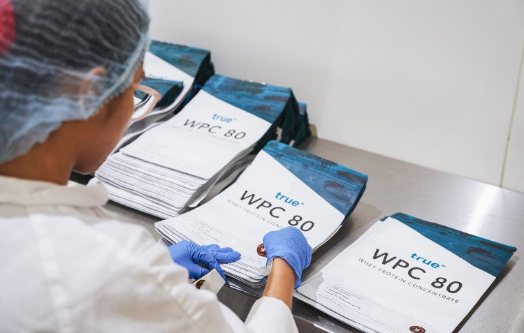 A True Protein team member in a hygienic lab coat and gloves carefully inspects and stacks pouches of WPC 80 Whey Protein Concentrate, showcasing our commitment to quality control at our Australian production facility.