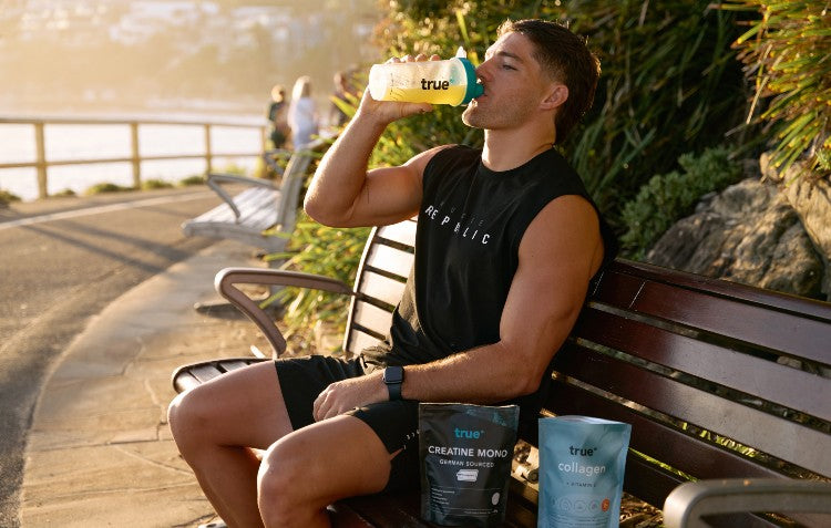 An athletic man in workout gear sits on a park bench along a sunny, coastal path, drinking from a yellow True Protein shaker. Next to him are pouches of True Protein Creatine Mono and Collagen supplements.